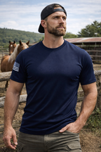 Man wearing a navy blue t-shirt with a patch, standing outdoors with horses in the background.