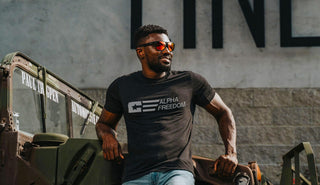 A man standing confidently in front of a hammer throw track, wearing a patriotic black t-shirt that reads "Alpha Freedom."