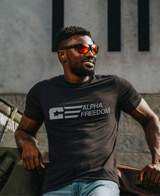 A man standing confidently in front of a hammer throw track, wearing a patriotic black t-shirt that reads "Alpha Freedom."